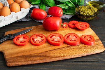 Sliced Roma Tomatoes on a Wooden Cutting Board: Slices of fresh plum tomatoes with eggs, basil, and a dish of pesto on the background