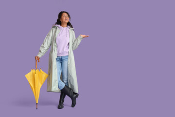 Happy young African-American woman in raincoat with umbrella on lilac background