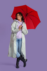 Happy young African-American woman in raincoat with umbrella on lilac background