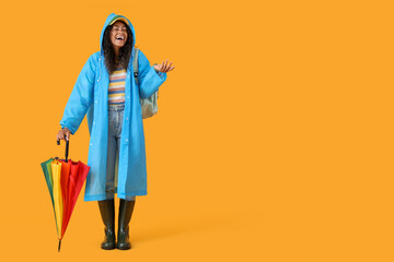 Happy young African-American woman in raincoat with umbrella on yellow background