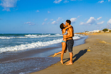 Happy young couple in bikini and shorts enjoying summer at the beach at dusk, breathing fresh air, kissing and teasing one another