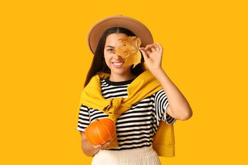 Stylish young woman with fresh pumpkin and autumn leaf on yellow background