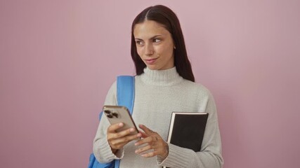 Woman using smartphone while holding book against pink background showing a beautiful young hispanic woman with brunette hair wearing casual attire over a pink isolated wall