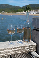 Bottle and glass of cold rose wine from Provence and wooden yacht boota pier on white sandy beach Plage de Pampelonne near Saint-Tropez, summer vacation in France