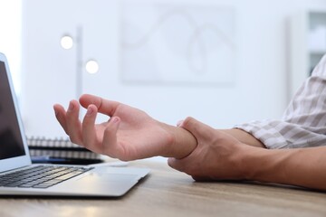 Man suffering from pain in wrist while working on laptop at table indoors, closeup. Carpal tunnel...