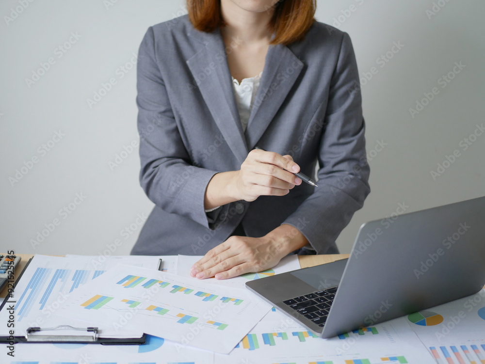 Wall mural young asian business woman sitting at a table holding a pen, calculating graphs and planning the pro