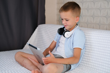Naklejka premium Boy plays games on tablet, lies on sofa. Laptop, home education. Knowledge website, educational software and small child broadcasting youth development lesson on headphones.
