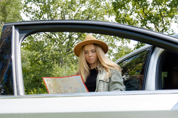 Smiling young woman in hat stoped on road to check the route on travel map. Local solo travel on weekends concept. Exited woman explore freedom outdoors in forest. Unity with nature lifestyle, rest