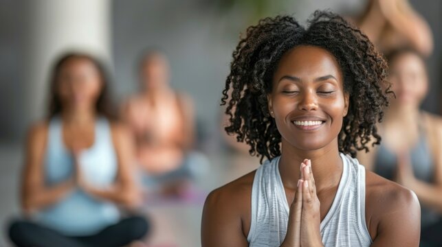 African woman with frizzy curly hair smiling warmly as she sits in a group yoga class. Hands put together in a prayer pose, creating a sense of calm, contentment, and mindfulness.
