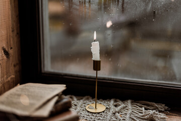 A candle stands on a wooden windowsill, raindrops outside the window, autumn mood, an open book © ShvedKristina