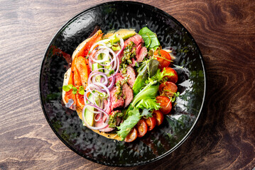 A vibrant dish featuring sliced tomatoes, onions, greens, and seasoned meat on a dark plate against a wooden background. Perfect for culinary themes, food blogs, or restaurant promotions.