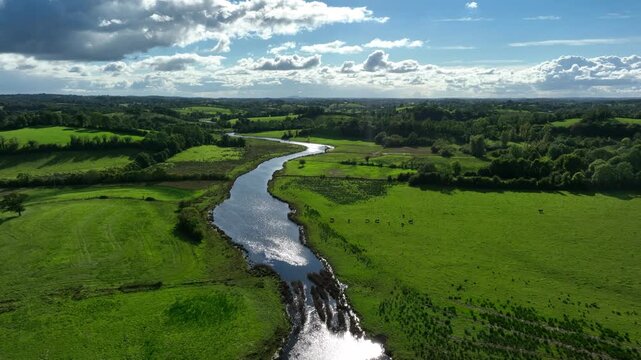 Finn River, County Monaghan, Ireland, September 2022. Drone ascends and pulls backwards with sunlight glistening in calm river water with panoramic views of grass and pasture.