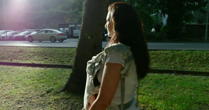 Woman walking with baby in a carrier on a sunny day, facing the camera, side profile view near a tree with blurred background, cars passing by, outdoor urban setting, summer light