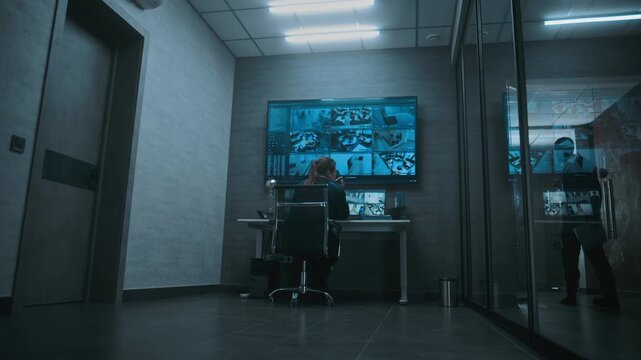Female surveillance operator controls security cameras using computer, talking on walkie talkie. CCTV footage on multiple monitors and big digital screen. African American worker walks by. Back view.