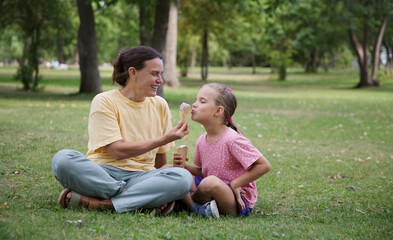 Happy family mom and child daughter eats ice cream having fun and doing picnic outdoor at city park on the green grass in summer day. Happy childhood. copy space