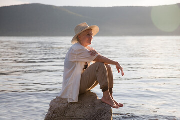 smiling elderly woman wearing a straw hat enjoys the sun sitting on a stone on the seashore or...