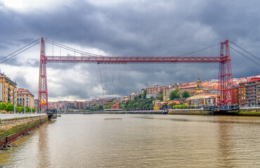 The Vizcaya Bridge, a large red transporter bridge spans across a river, with an overcast sky and buildings lining the riverbanks. The structure connects the towns of Portugalete and Las Arenas.