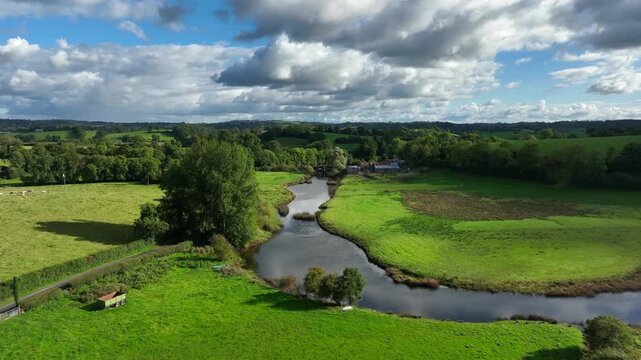 Finn River, County Monaghan, Ireland, September 2022. Drone pushes forward above river over bridge passing rusted farm house nestled between trees on cloudy day.