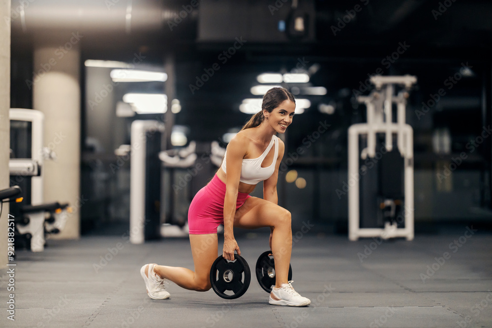 Wall mural happy strong sportswoman in trendy sportswear doing lunges with weight plates in a gym.