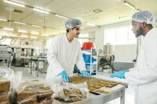 Two multicultural food factory workers in sterile uniforms collecting fresh biscuits and putting them into a bags.