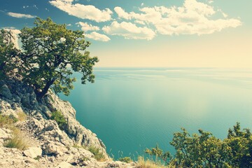 Naklejka premium Serene landscape featuring a rocky cliff, lush vegetation, and tranquil waters under a bright sky with fluffy clouds.