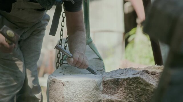 Professional craftsman in workwear and respirator carving stone with hammer and chisel while working outdoors