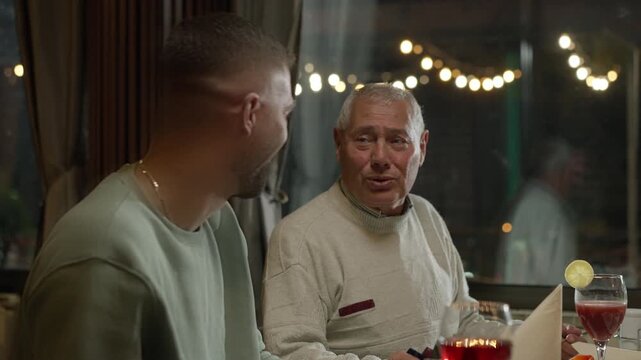 Senior Caucasian Man Talking Jokes to His Son at the Restaurant. Glasses of Juice on the Table. Concept of Uncle and Nephew, Humor, Laughing, Bonding, Funny, Relaxation, Enjoyment, Happiness