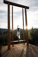 A woman in a white dress dances and poses on a swing high in the mountains at sunset. Freedom concept