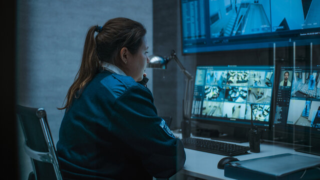 Female surveillance operator controls security cameras with face recognition in monitoring center using computer, talks on landline phone. CCTV footage on multiple monitors and big digital screen.