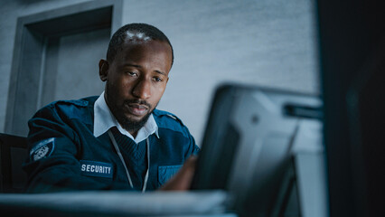 African American male security operator or dispatch in uniform uses tablet computer, looks at...