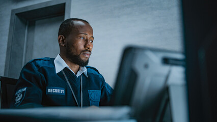 African American male security operator or dispatch in uniform uses tablet computer, looks at...