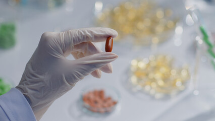 The researcher's hand is holding a multivitamin pill. The blurred background below are laboratory dishes containing various drugs. Scientific research on drugs and functional foods