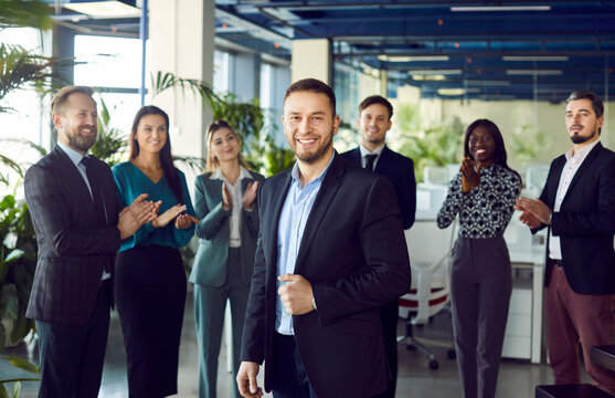 Smiling young attractive boss and business leader man proudly looking at camera with team of people applauding in background in office. Business team or group of staff congratulating their leader.