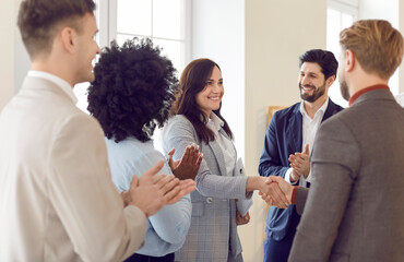Woman shaking hands with a colleague during a meeting, surrounded by a team or group of applauding colleagues. People celebrating agreement or deal, successful and collaborative business environment.