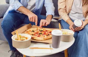 Delivery or takeaway food. Young couple enjoying lunch at home with delivered pizza, salad and hot coffee. Cropped image of couple sitting on sofa and enjoying food ordered from delivery service.