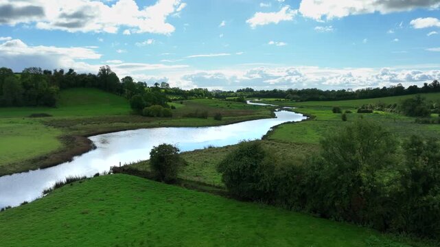 Finn River, County Monaghan, Ireland, September 2022. Drone pushes forward above trees growing on fence line with water blowing ripples over water reflecting cloudy sky as sheep graze in pasture.
