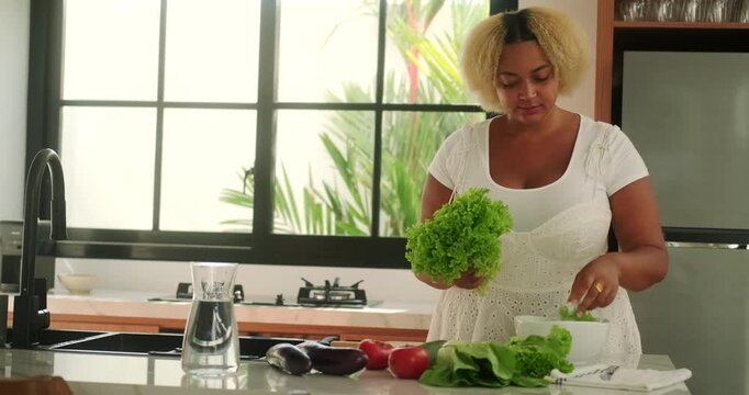 Young African American curvy woman standing in the kitchen preparing a salad of green lettuce leaves, different vegetables on the table. Diet and healthy lifestyle. Body positive.