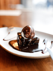 delicious dessert with black sponge cake, covered in chocolate cream, and white ice cream, served on a beautiful white plate on a wooden table in a cozy, charming restaurant