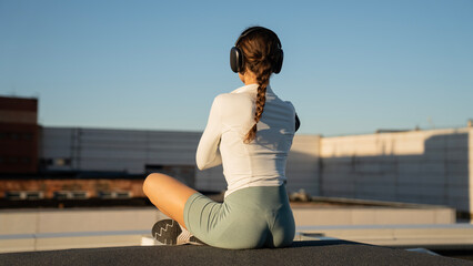Fototapeta premium Young athletic girl sitting on concrete block on roof and meditating against buildings in city