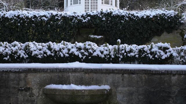 Romantic white garden house on a snowy winter day. Snow covered steps leading to a little cottage in a city park. There are many evergreen bushes and deciduous trees in the fore- and background.