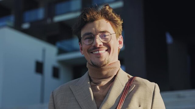 Smiling happy man with glasses looks at the camera standing in the sunlight city. Businessman. Stylish guy. Slow motion