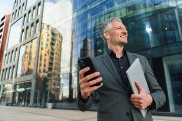 A confident businessman in a gray suit holds a smartphone and a tablet, smiling while looking up in a modern urban setting.