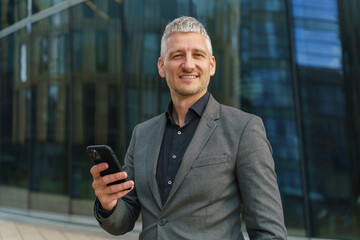 A businessman in a gray suit smiles while holding his phone, standing confidently in front of a modern glass building.