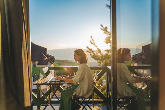 Young woman sitting on balcony with view of mountains and sunset working using laptop