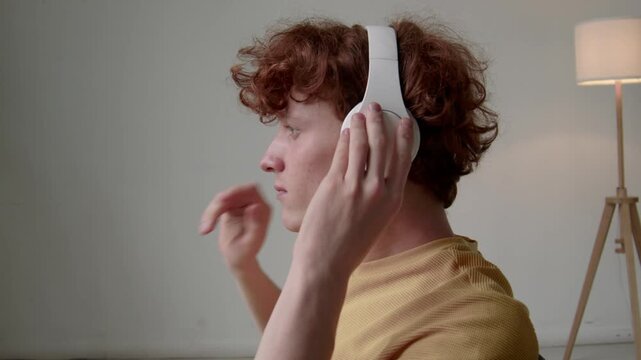 Side closeup of gen Z Caucasian guy with red curly hair putting on white wireless headset and listening to music or podcast at home