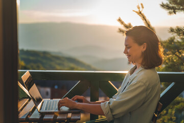 Young woman sitting on balcony with view of mountains and sunset working using laptop