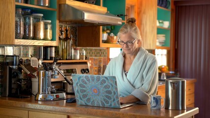 Happy mature woman in bathrobe telecommuting working on her laptop in her kitchen.