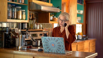 Smiling happy mature woman talks on phone while looking at computer screen in a kitchen.