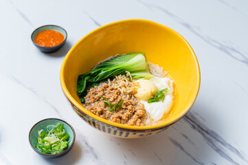 A vibrant yellow bowl of Asian noodles topped with minced pork, poached egg, and bok choy, served with green onions and chili sauce on a white marble surface, ideal for food photography