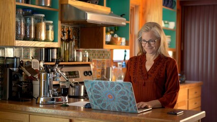 Smiling happy mature woman with glass of wine working on a laptop on the kitchen counter.